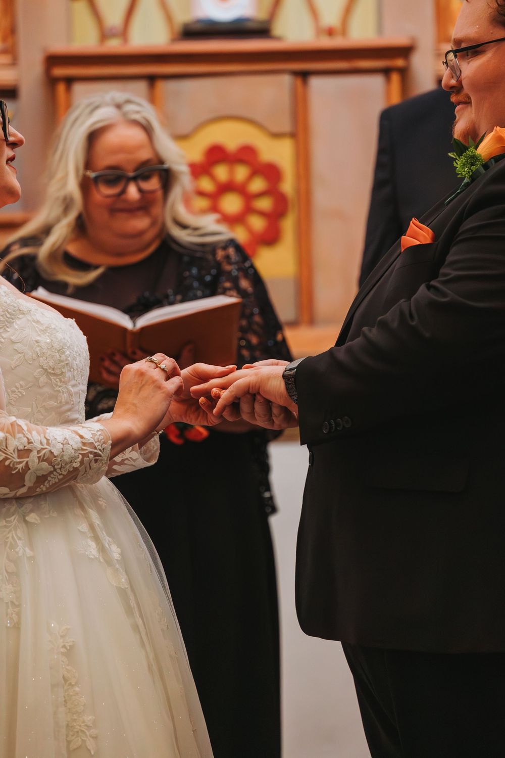 Bride placing wedding ring on partner's finger during ceremony, officiant holding book in background.