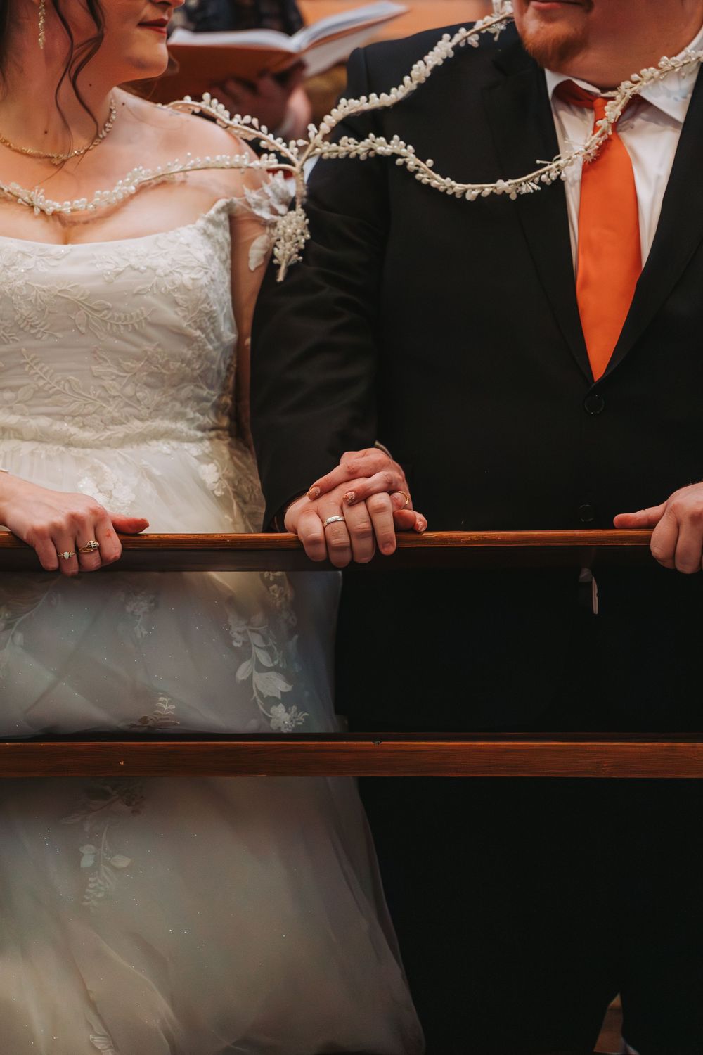 Bride and groom holding a wooden rail during wedding ceremony, bride in lace dress, groom in black suit with orange tie.