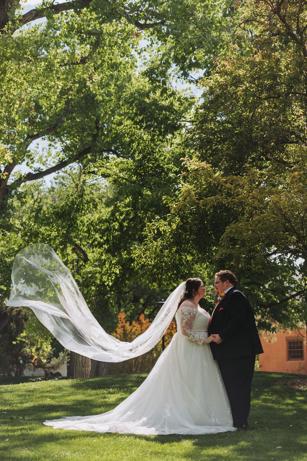 Bride and groom share a romantic moment outdoors, with her flowing veil caught by the wind beneath a large green tree.