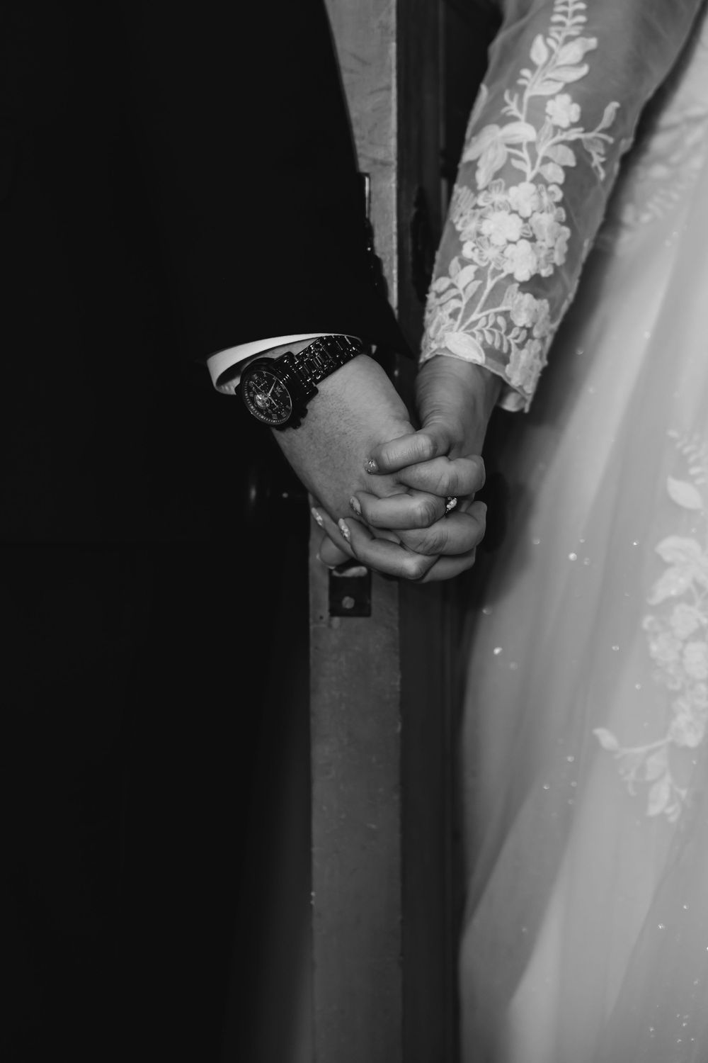 Black and white photo of bride and groom holding hands, showing wedding ring and watch, with lace dress detail.