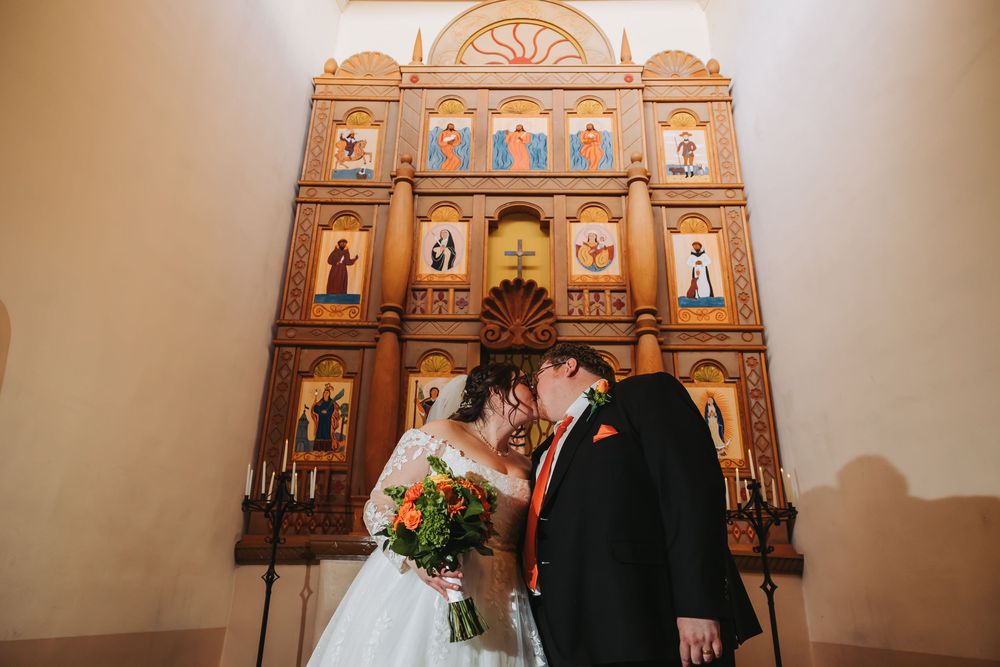 Bride and groom kissing in front of ornate wooden Orthodox iconostasis during wedding ceremony in a church.