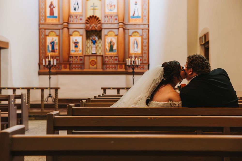 Bride and groom sharing an intimate kiss in a church pew before a ornate wooden altar with candles.