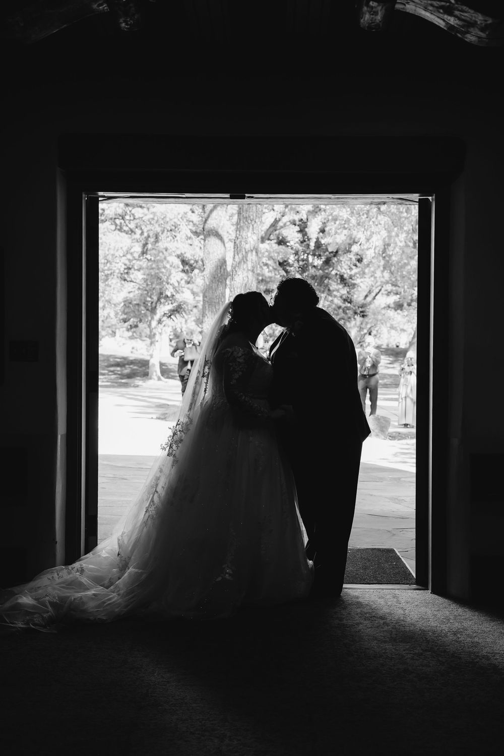 Black and white silhouette of bride and groom kissing in church doorway with natural light and trees behind them.