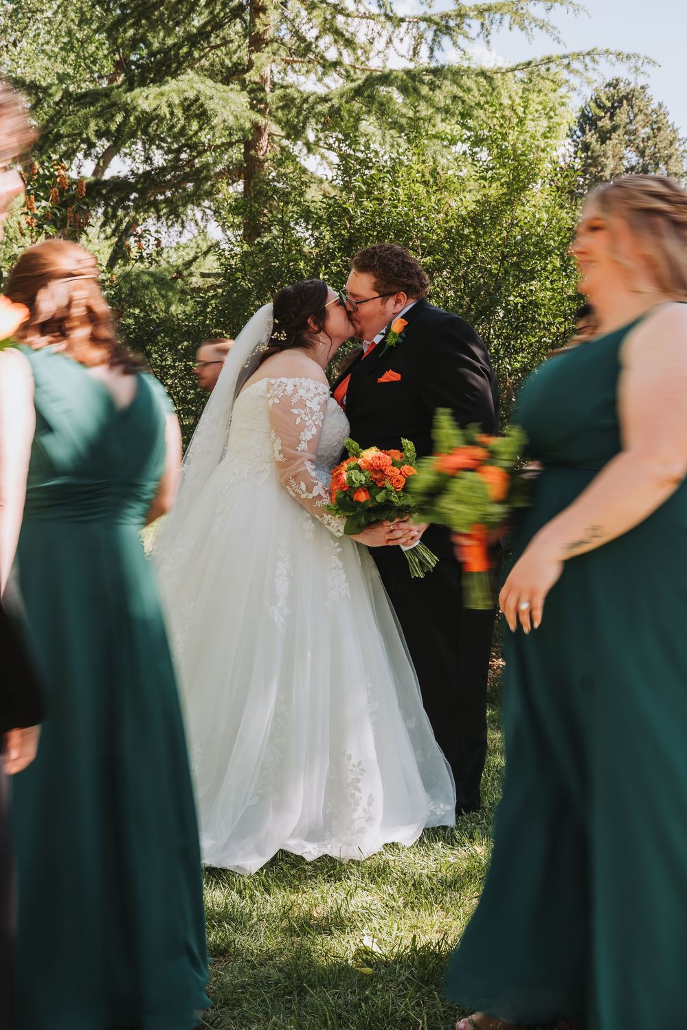 Bride and groom share first kiss at outdoor wedding ceremony, surrounded by bridesmaids in emerald green dresses.