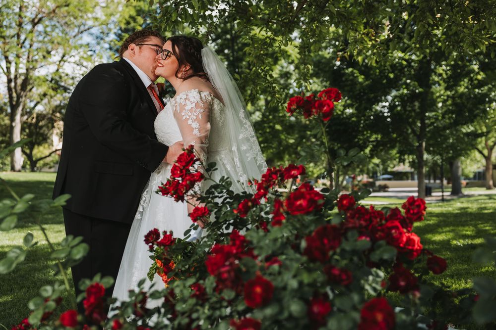 A bride and groom share a kiss surrounded by lush green trees and vibrant red roses in an outdoor park setting.
