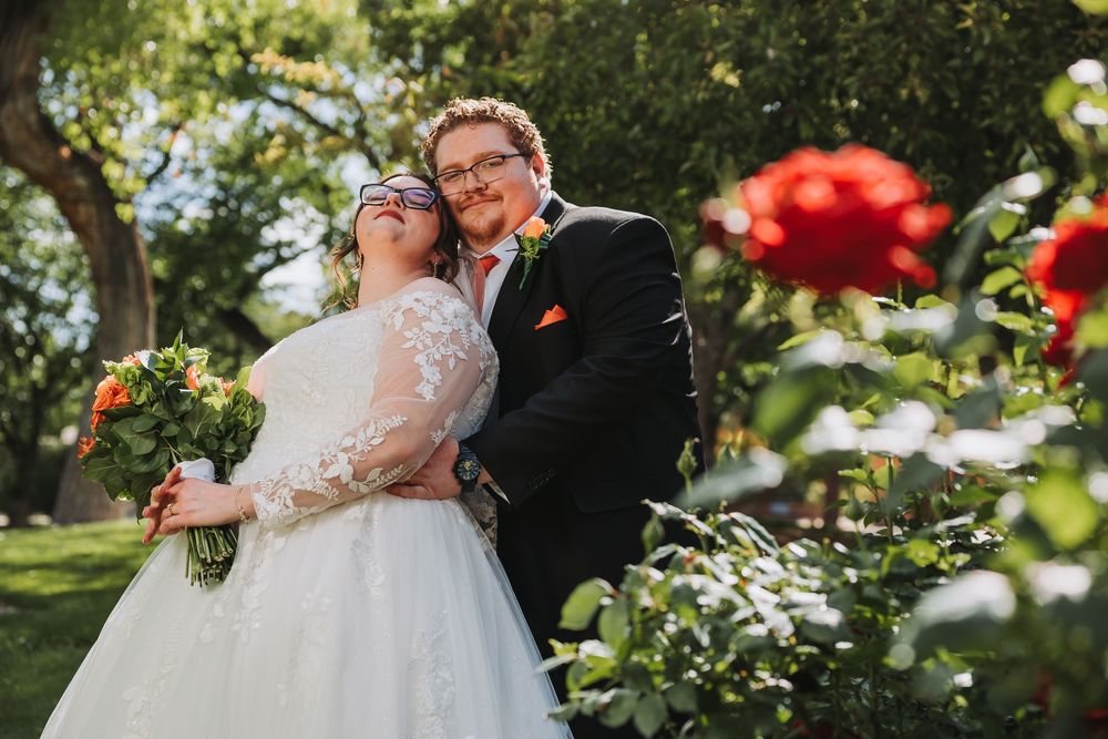 Bride and groom posing together in a lush garden with red roses, bride holding a colorful bouquet in wedding attire.
