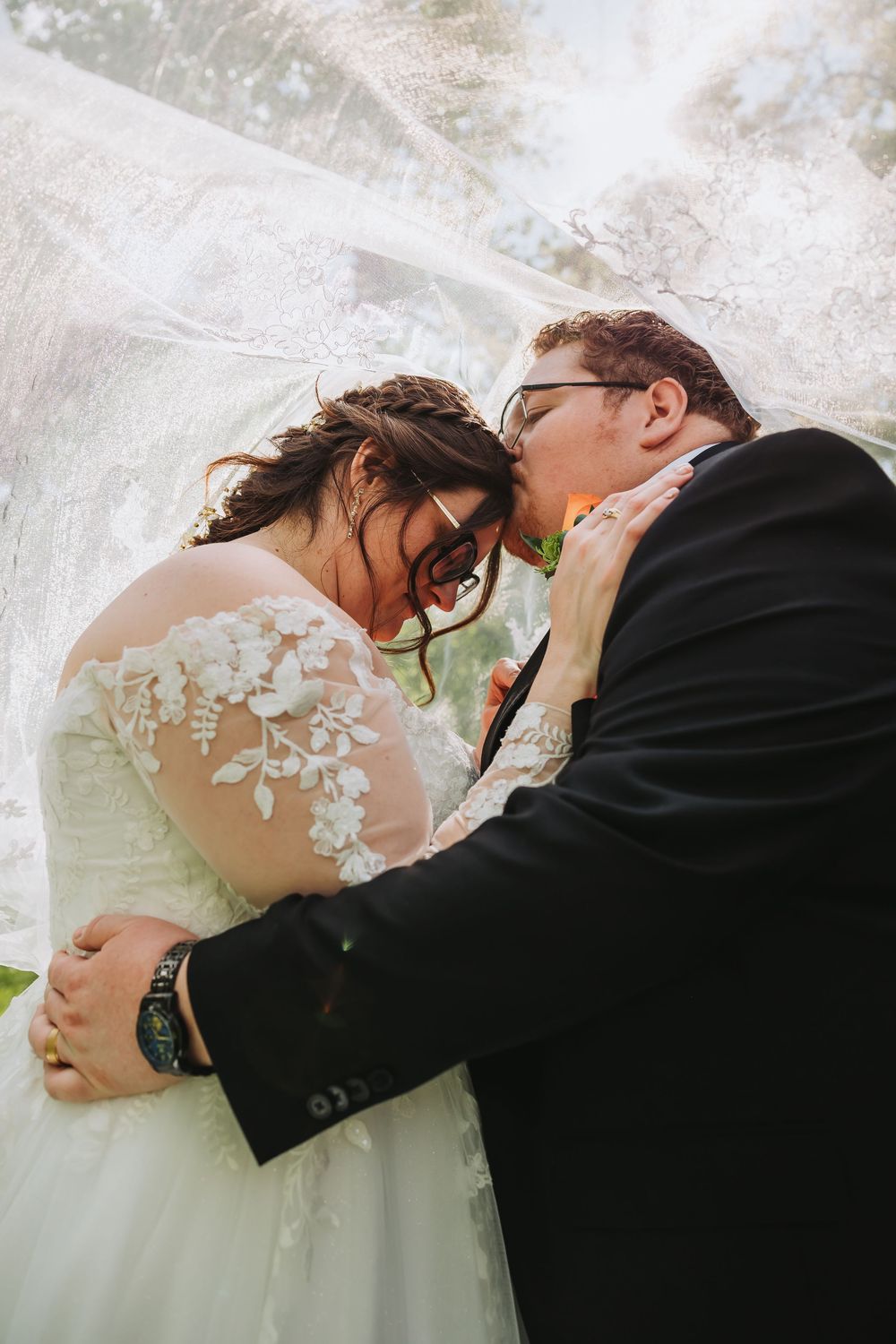 Bride and groom sharing a tender moment under flowing white veil, bride in lace dress, groom in black suit.