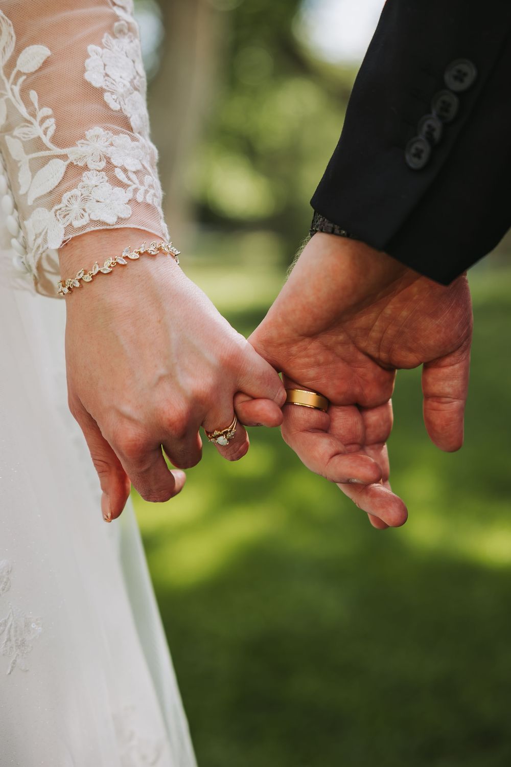 Newlywed couple holding pinkies, wearing gold wedding rings, bride in lace dress with diamond bracelet.