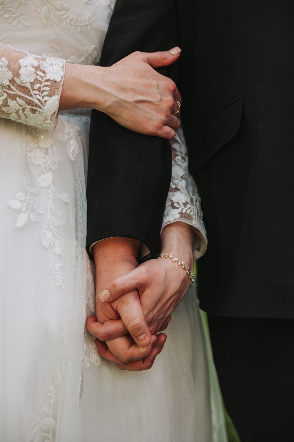 Bride and groom holding hands, bride in lace wedding dress with bracelet, groom in black suit, romantic wedding moment.