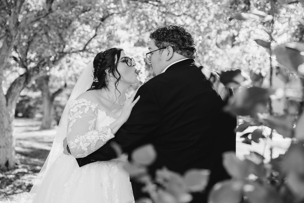 Black and white photo of a bride and groom sharing a kiss outdoors under trees during their wedding ceremony.