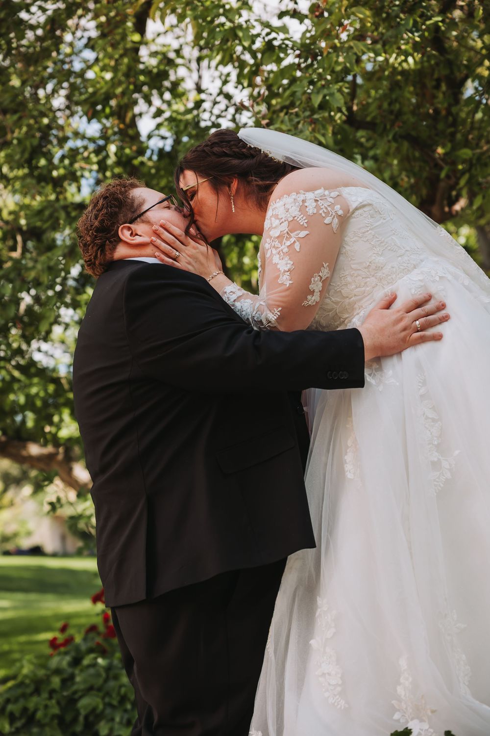 Two brides share a first kiss in a lush garden, one in a lace wedding gown with veil, the other in a black suit.