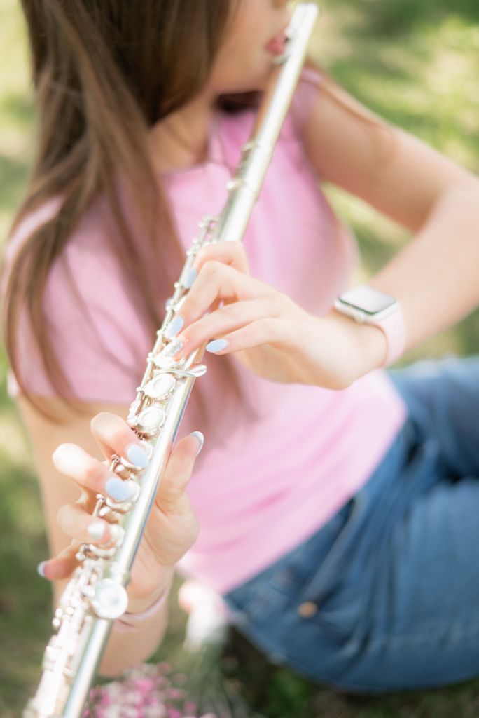 Young girl in pink shirt playing silver flute outdoors in a park setting.