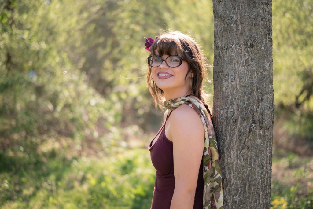 A smiling woman with glasses and a flower in her hair leans against a tree in a sunlit forest wearing a burgundy dress.