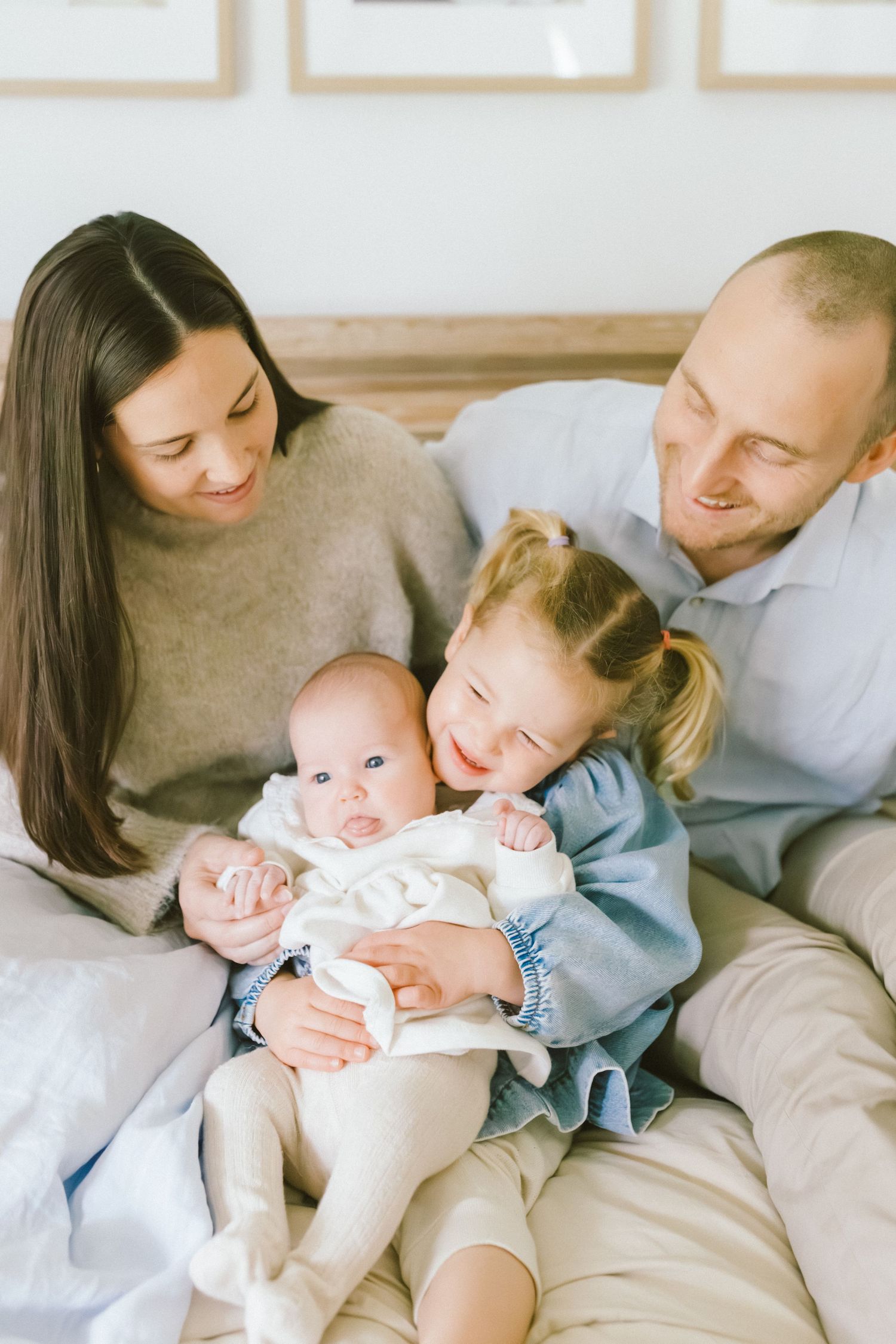 A happy family of four sits together on a bed, with parents smiling at their toddler daughter holding a newborn baby.