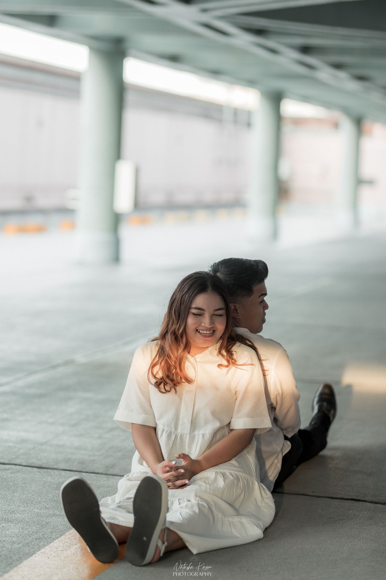 A couple sits on the floor under a covered structure, the woman in white smiling warmly as her partner leans close behind her.