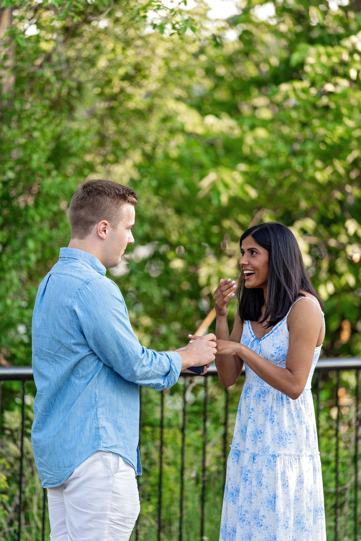 Joyful couple holding hands and laughing together during a marriage proposal in a green park.