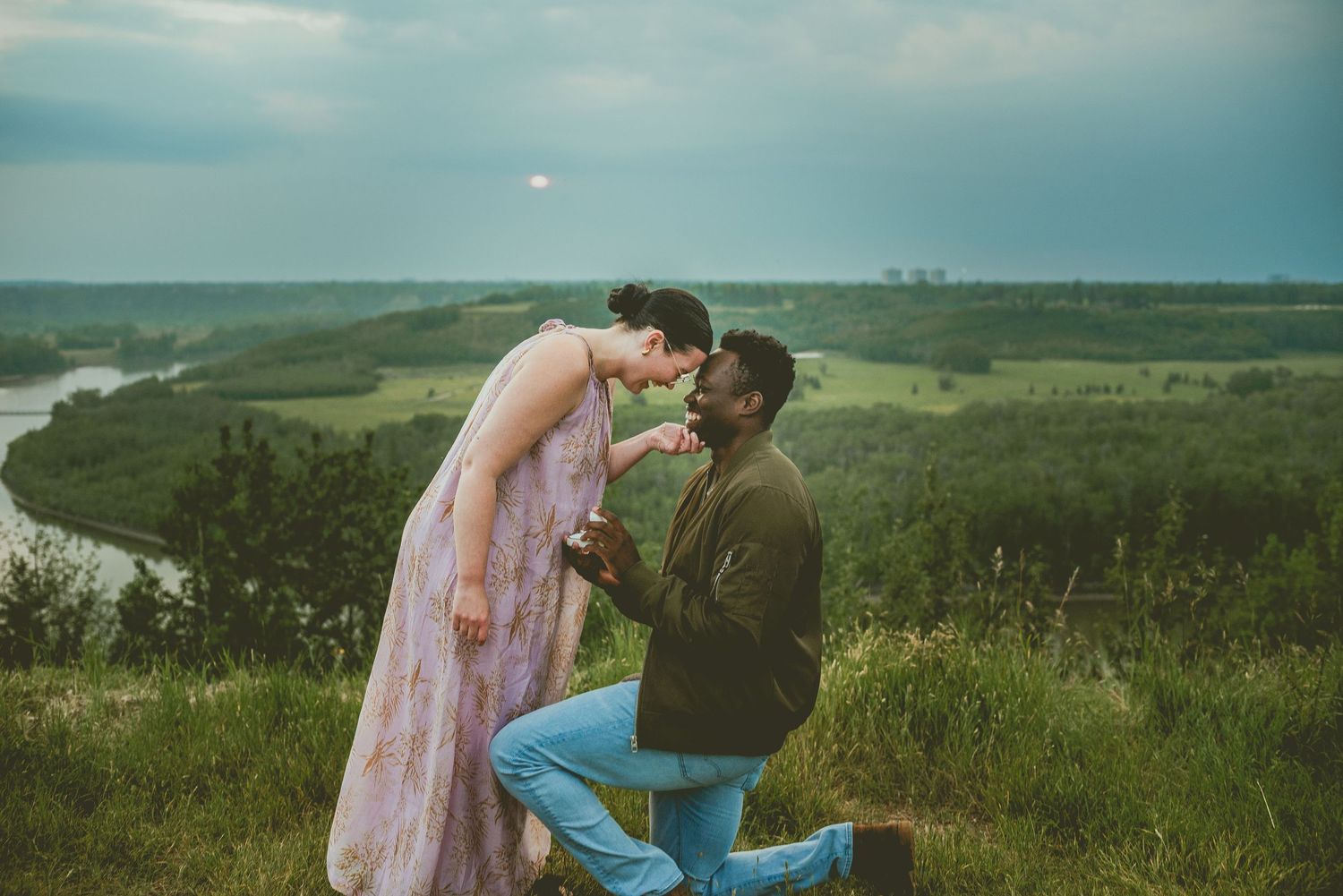 Romantic interracial couple sharing a proposal moment on a scenic hilltop overlooking a river valley at dusk.