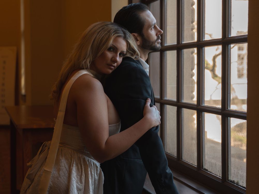 Couple embracing by a window in warm golden light, woman in cream dress with shoulder bag hugging man in dark suit.