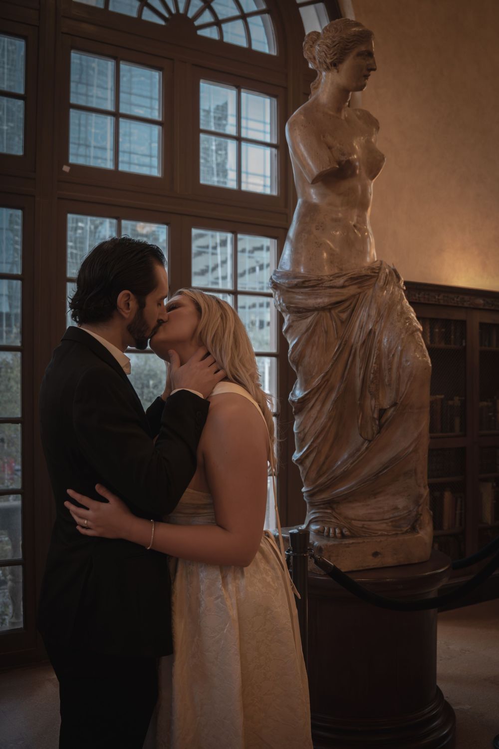Couple sharing a romantic kiss near a classical Venus de Milo statue in an elegant dimly lit museum.