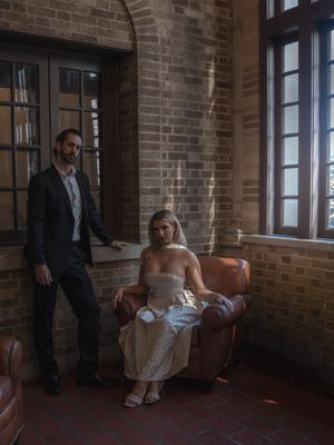 Bride in white dress seated in leather chair with groom standing beside her in brick-walled venue.