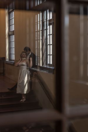 Wedding couple embracing near tall windows on a sunlit staircase, captured through a glass reflection.