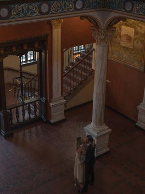 Aerial view of wedding couple embracing between marble columns in a grand historic building lobby.