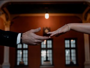 Close-up of bride and groom reaching toward each other with fingertips touching in ornate venue.