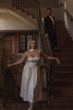 Bride in strapless white dress poses on staircase with groom descending behind her in elegant venue.