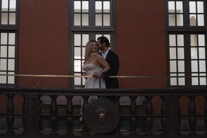Wedding couple embracing on an ornate balcony with large windows and decorative railing behind them.