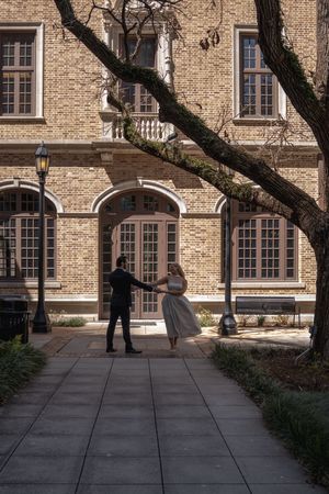 Couple dancing on a brick pathway outside a historic brick building with arched windows and a bare tree.