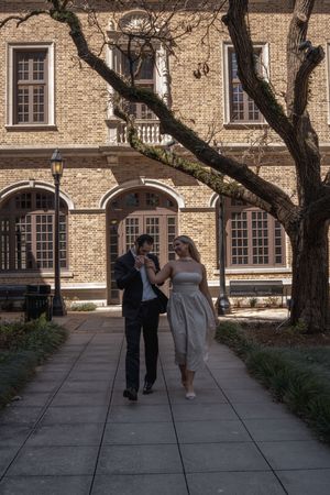 Romantic couple walking hand-in-hand along a sunlit pathway beside a classic brick building with arched windows.