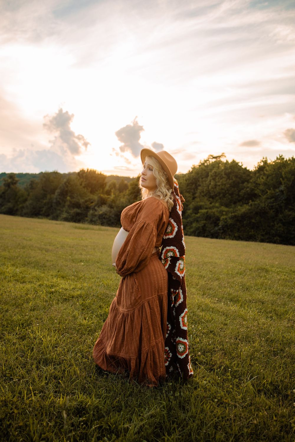 Pregnant woman in rust boho dress and hat stands in green field at golden sunset.