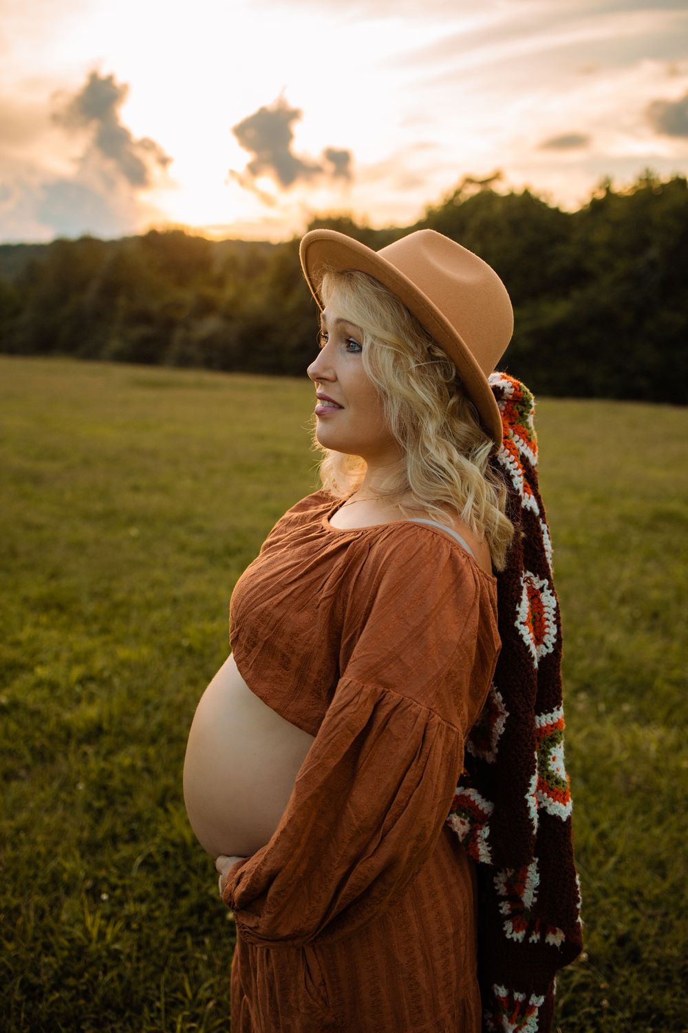 Pregnant woman in rust dress and tan hat holds belly at golden hour sunset in green field.