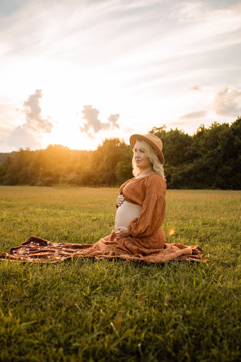 Pregnant woman in rust dress and hat sits on blanket in sunny green field at golden hour sunset.