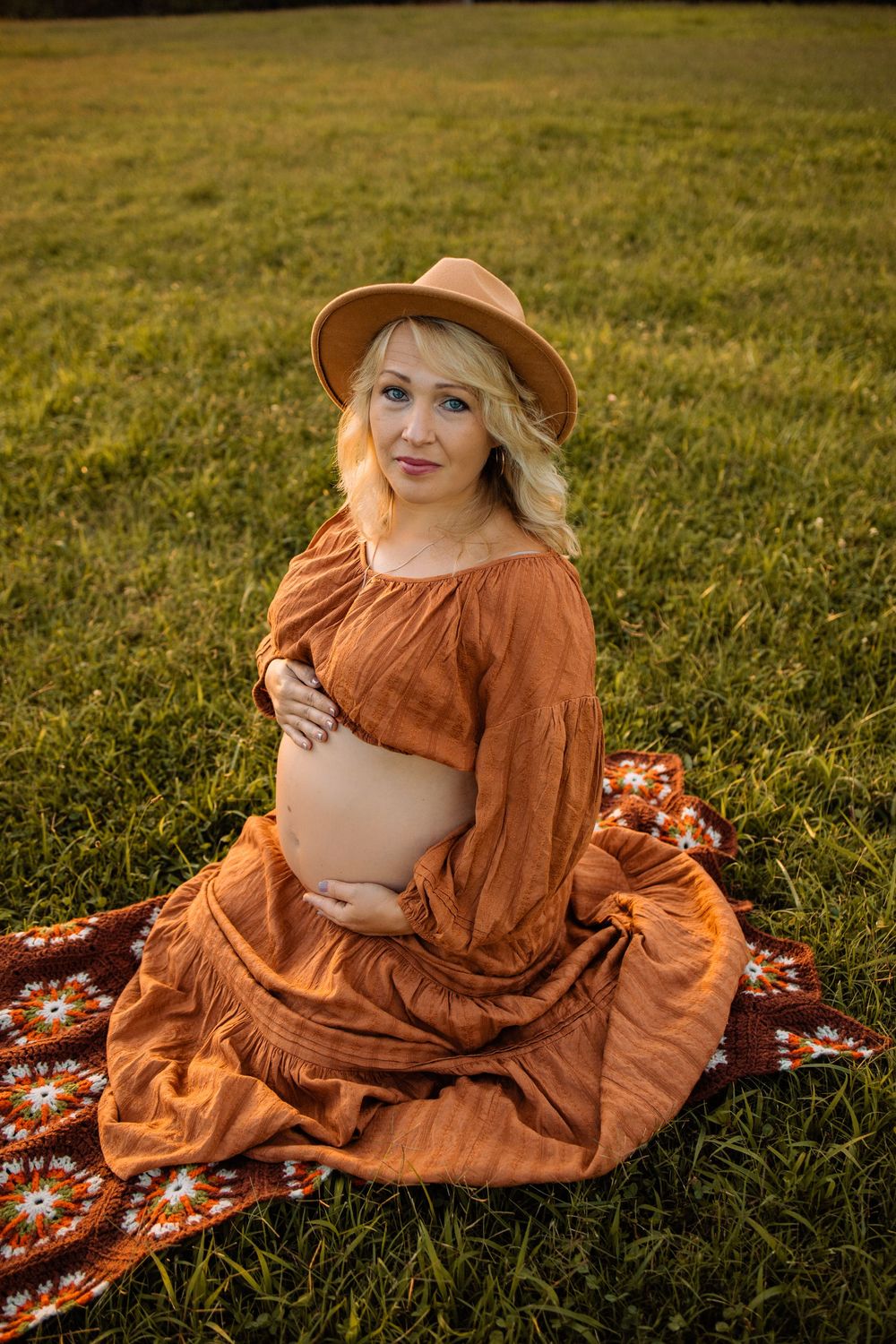 Pregnant blonde woman in rust dress and hat sits on floral blanket in grassy field.