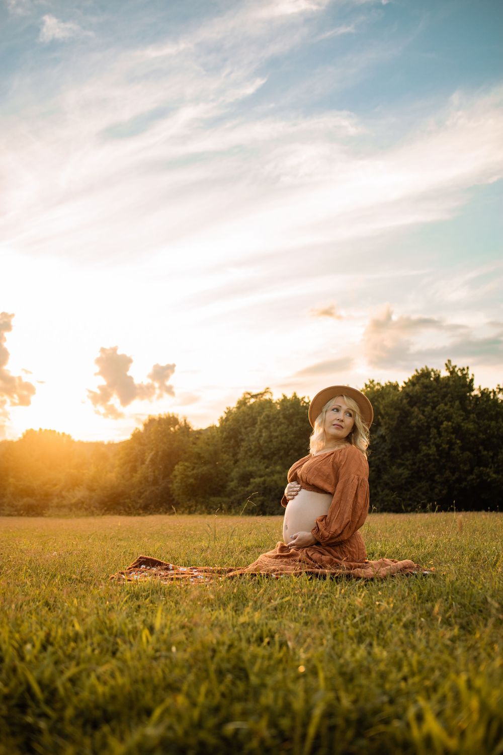 Pregnant woman in brown dress and hat sitting on grass field at golden sunset, cradling baby bump outdoors.