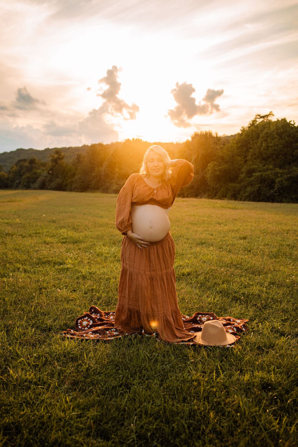 Pregnant woman in rust dress holds baby bump at golden hour sunset in lush green field.