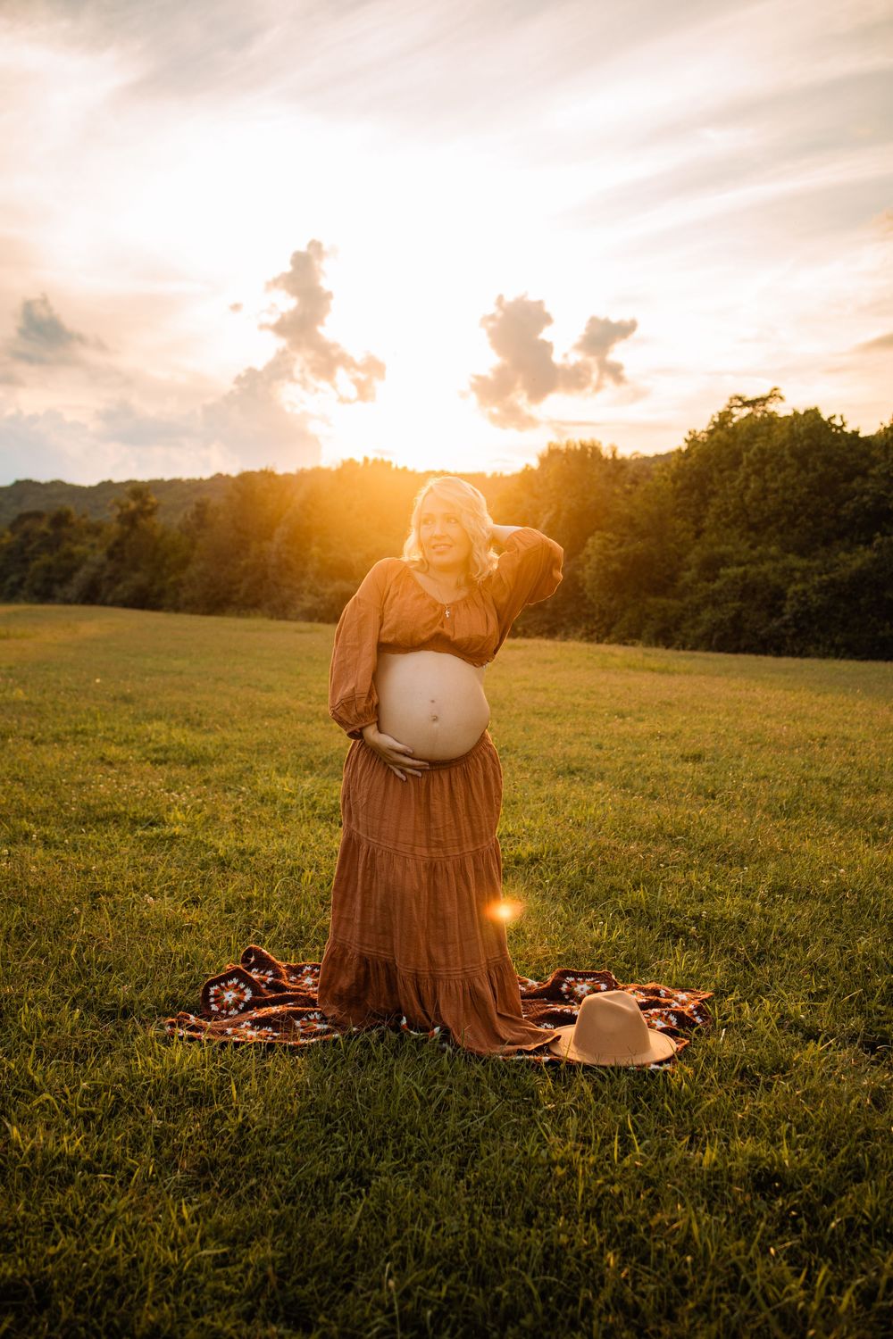Glowing pregnant woman in amber dress cradles belly at golden hour in green meadow.