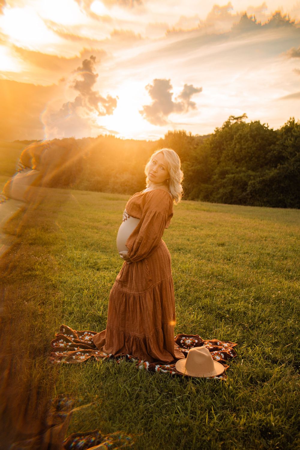 Pregnant woman in rust boho dress holding belly at golden sunset in green field with hat on blanket.
