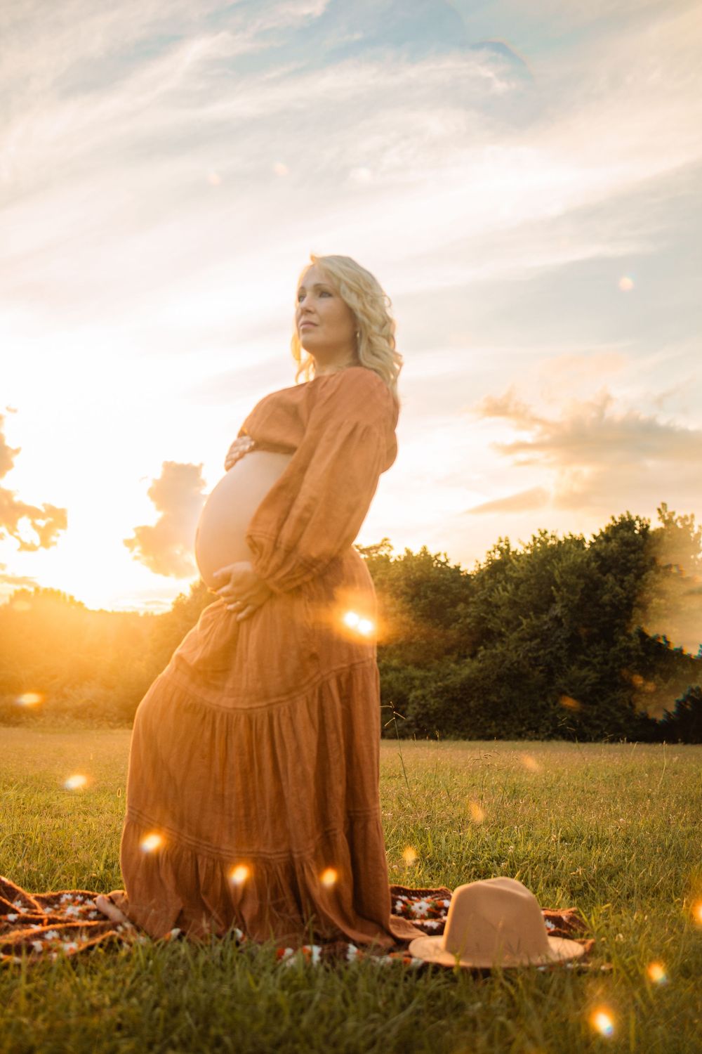Glowing maternity photo of blonde woman in rust boho dress at sunset with bokeh light effects.