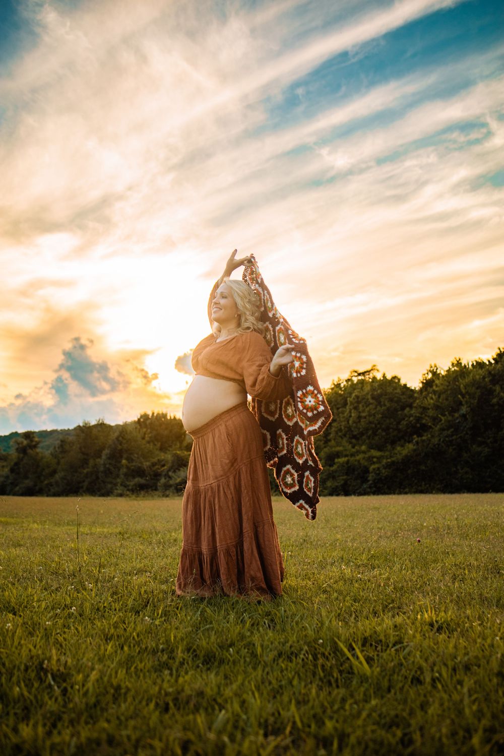 Pregnant woman in brown boho dress holding crochet shawl in sunlit field at golden hour sunset.