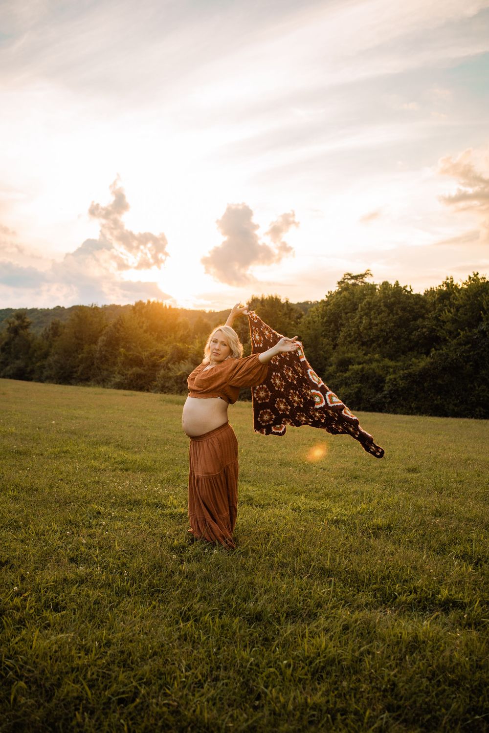 Pregnant woman in rust skirt twirls floral fabric at golden sunset in open green meadow.