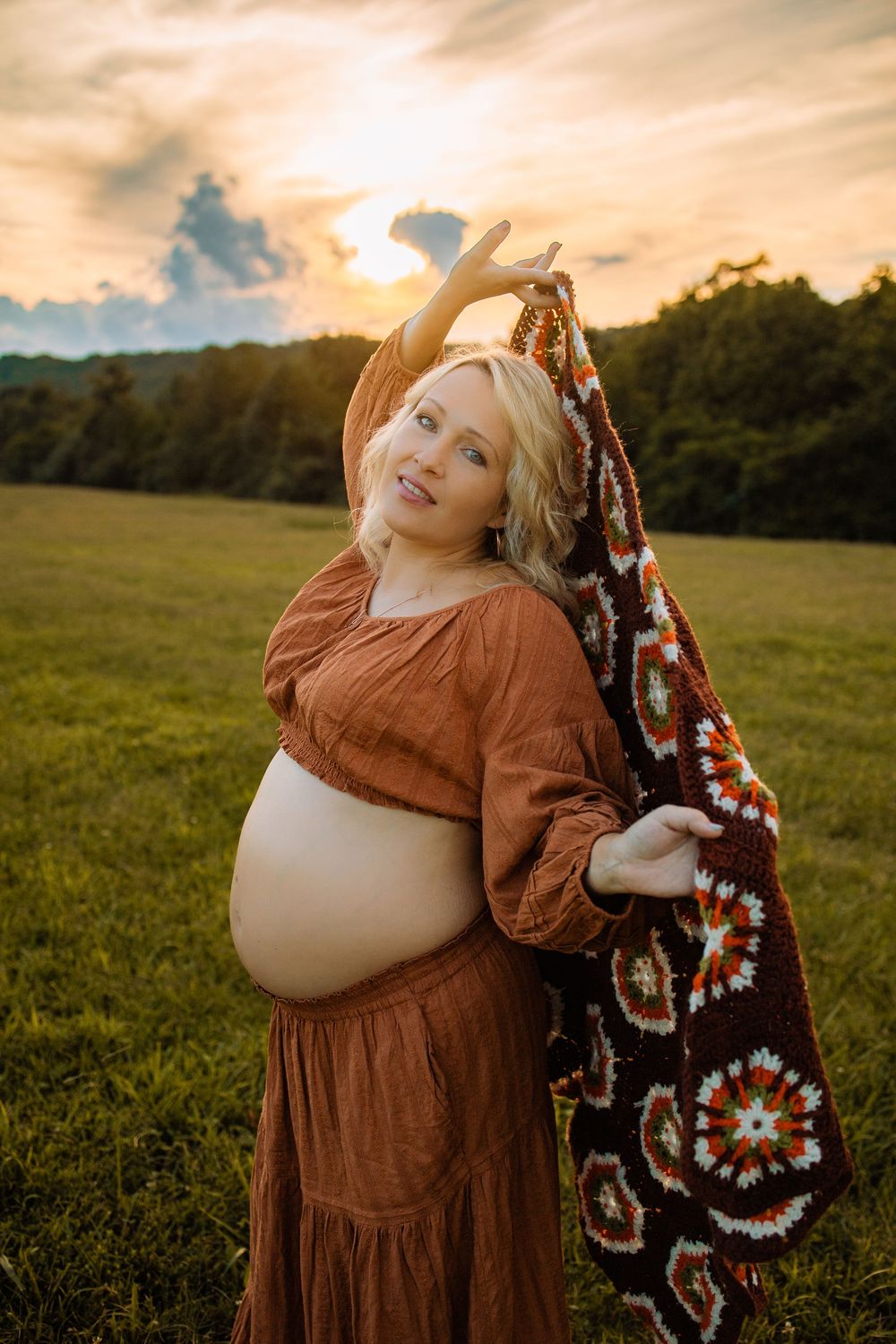 Pregnant woman in rust boho dress holds crochet blanket in golden sunset field.