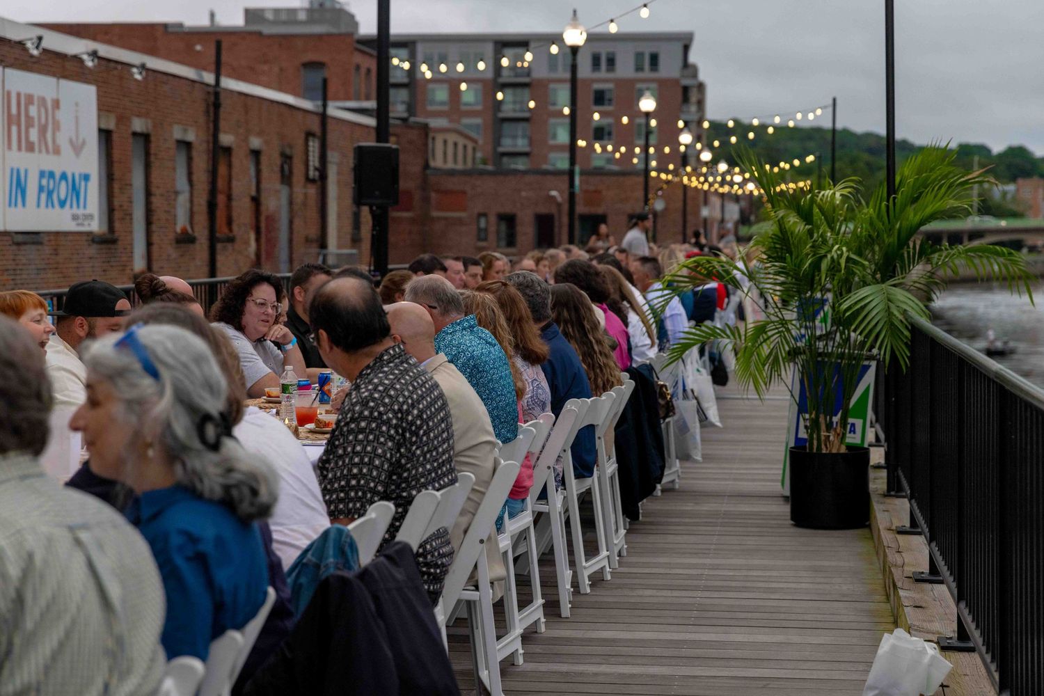 Outdoor waterfront dinner event with guests seated at long tables on a dock, string lights overhead, urban backdrop.
