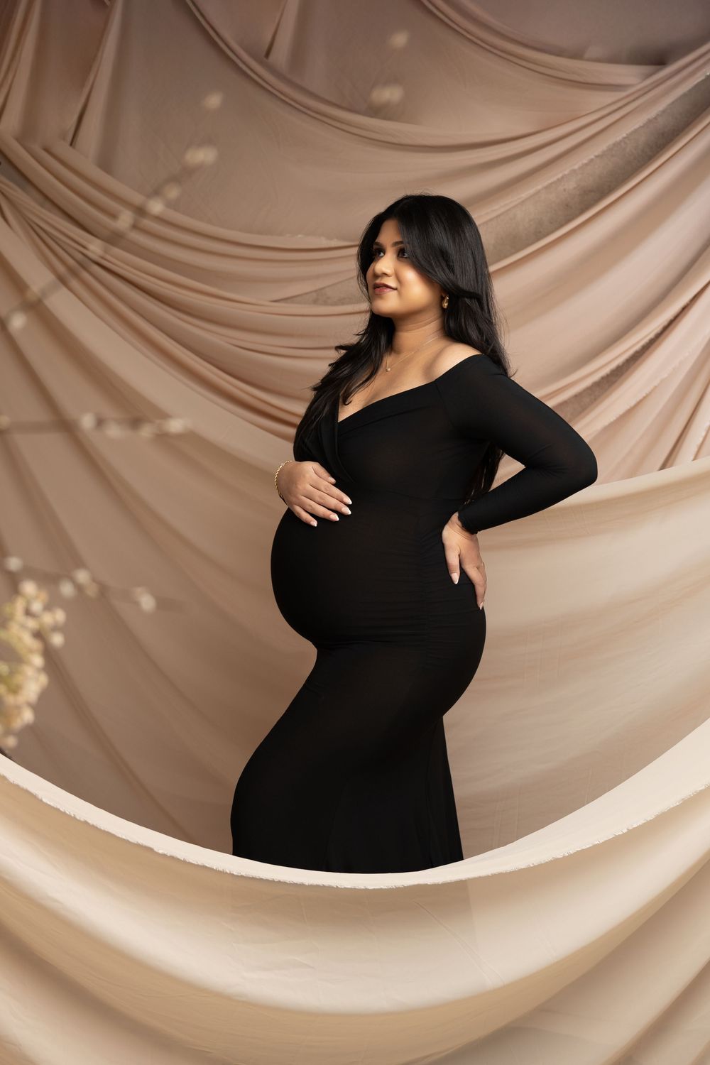 Pregnant woman in black form-fitting maternity gown poses gracefully against flowing beige fabric backdrop.