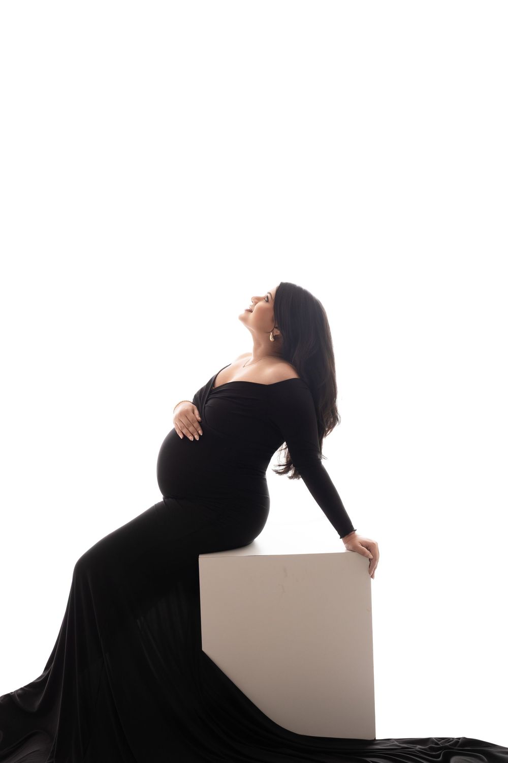 Pregnant woman in elegant black gown posing gracefully on white cube against white background.