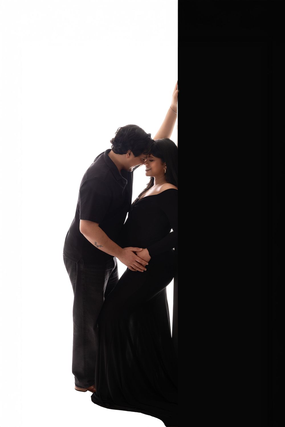 A couple in black clothing share an intimate moment, the pregnant woman cradling her bump against a bold black and white backdrop.