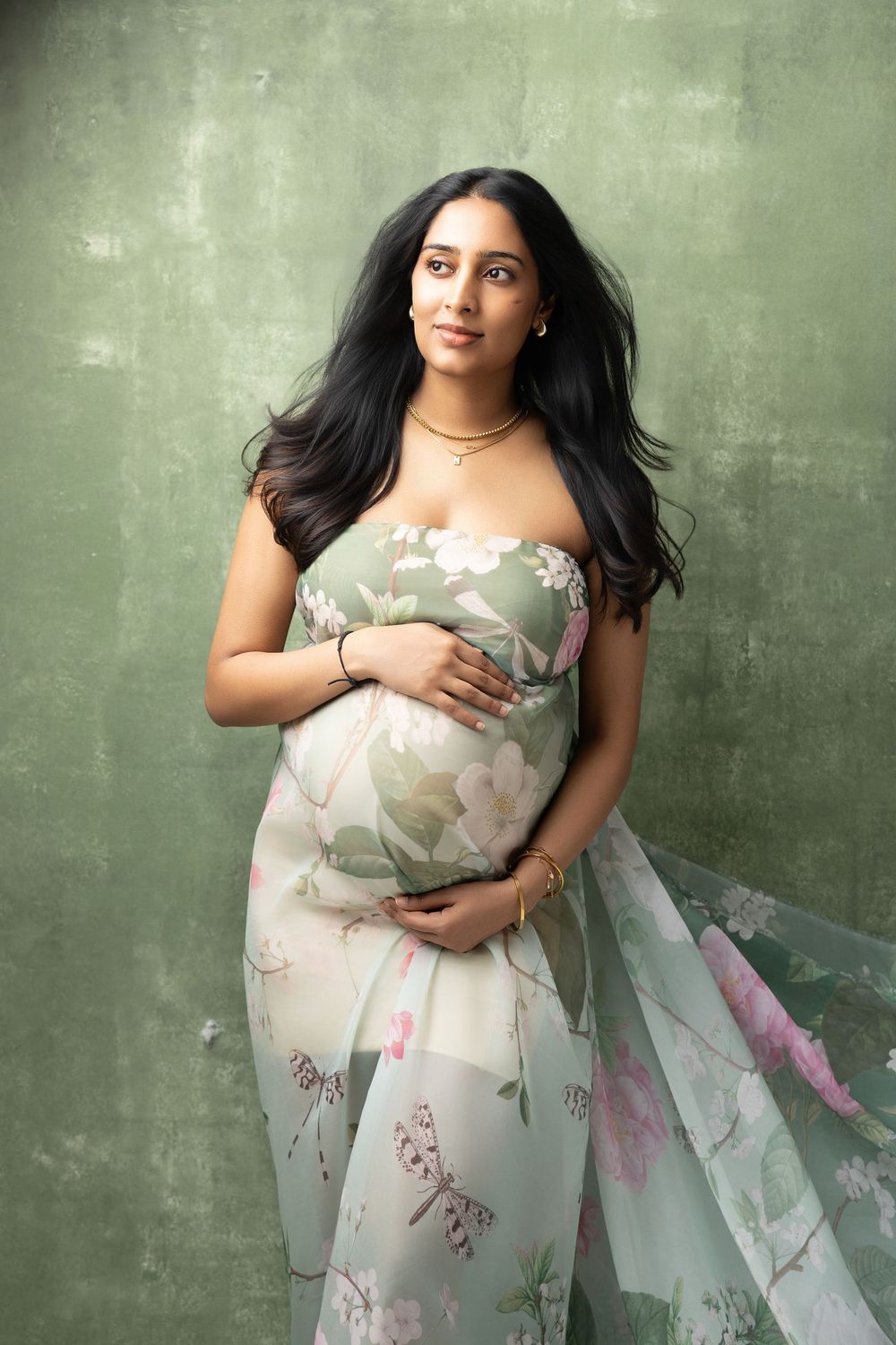 Pregnant woman in floral sage green gown cradling baby bump during elegant maternity portrait session.