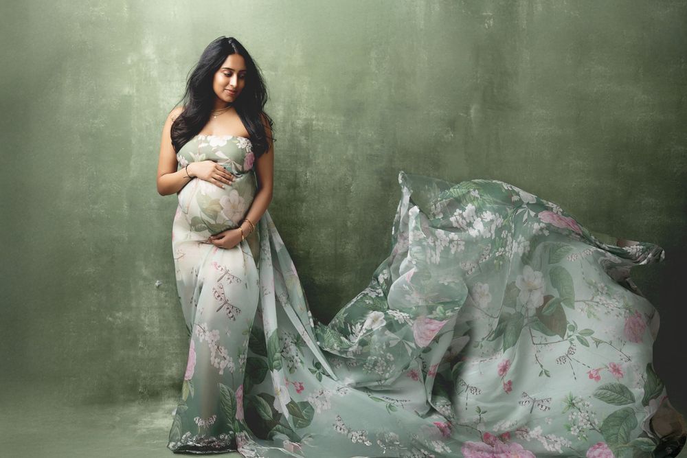 Pregnant woman in floral green maternity gown with flowing fabric, posing gracefully in a professional studio photo.