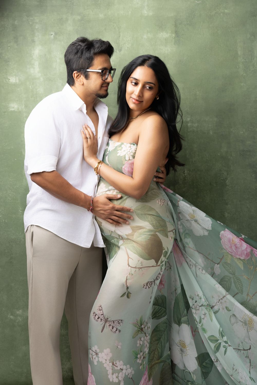 Pregnant Indian couple posing together in studio, woman in floral saree gown, man in white shirt, green background.